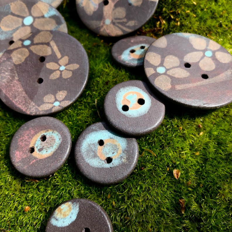 May include: A collection of dark gray ceramic buttons with blue and white floral designs. The buttons are arranged on a bed of green moss.