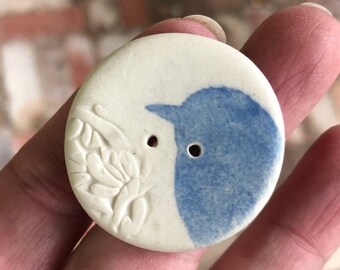 以下が含まれることがあります： 青い鳥のデザインが施された白いセラミックボタン。鳥は右を向いており、小さなくちばしがあります。ボタンには縫い付けるための穴が2つあります。