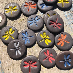Pode incluir: Uma coleção de azulejos redondos de cerâmica preta com desenhos de libélulas coloridas. Cada azulejo apresenta uma libélula diferente com várias cores, incluindo amarelo, vermelho, azul e laranja.
