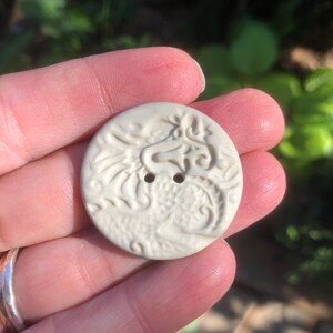May include: A white ceramic button featuring a raised design of a dragon. The button has two holes for sewing.