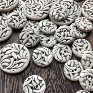 May include: A collection of white ceramic buttons with a black floral design. The buttons are various sizes and shapes, and some have two holes.