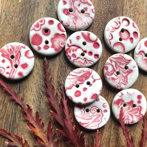 Può includere: Una collezione di dieci bottoni in ceramica bianca con un motivo floreale rosso. I bottoni sono rotondi e hanno due fori.