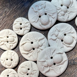 Peut inclure: Dix boutons en céramique blanche avec un motif d'abeille en relief. Les boutons sont de tailles et de formes différentes, mais ils ont tous un motif d'abeille similaire.