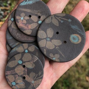 May include: A set of six brown ceramic buttons with a floral pattern in shades of brown, blue, and pink. The buttons have two holes each.