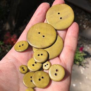 May include: A collection of 12 round, ceramic buttons in various sizes. The buttons are a muted yellow-green color with two holes each.