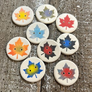May include: Ten round, off-white buttons with maple leaf designs in various colours, including red, orange, blue, and green. Each button has two holes for sewing. The buttons are arranged on a weathered wooden surface, showcasing a range of autumnal hues.