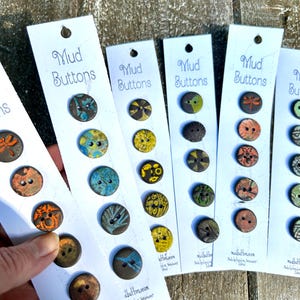 May include: Five cards with the text "Mud Buttons" on them. Each card has 10 round ceramic buttons with different designs and colors. The buttons are arranged in two rows of five. The cards are on a wooden surface.