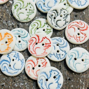May include: A selection of white, circular buttons featuring squirrel illustrations in shades of red, blue, green, and orange. Each button has two holes for sewing.