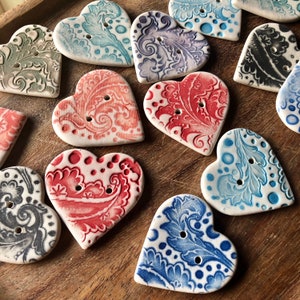 Peut inclure: Une collection de boutons en céramique en forme de cœur avec différents motifs floraux et paisley dans des tons de bleu, rouge et gris. Les boutons ont deux trous chacun et sont parfaits pour ajouter une touche de charme vintage à l'artisanat et aux projets de couture.
