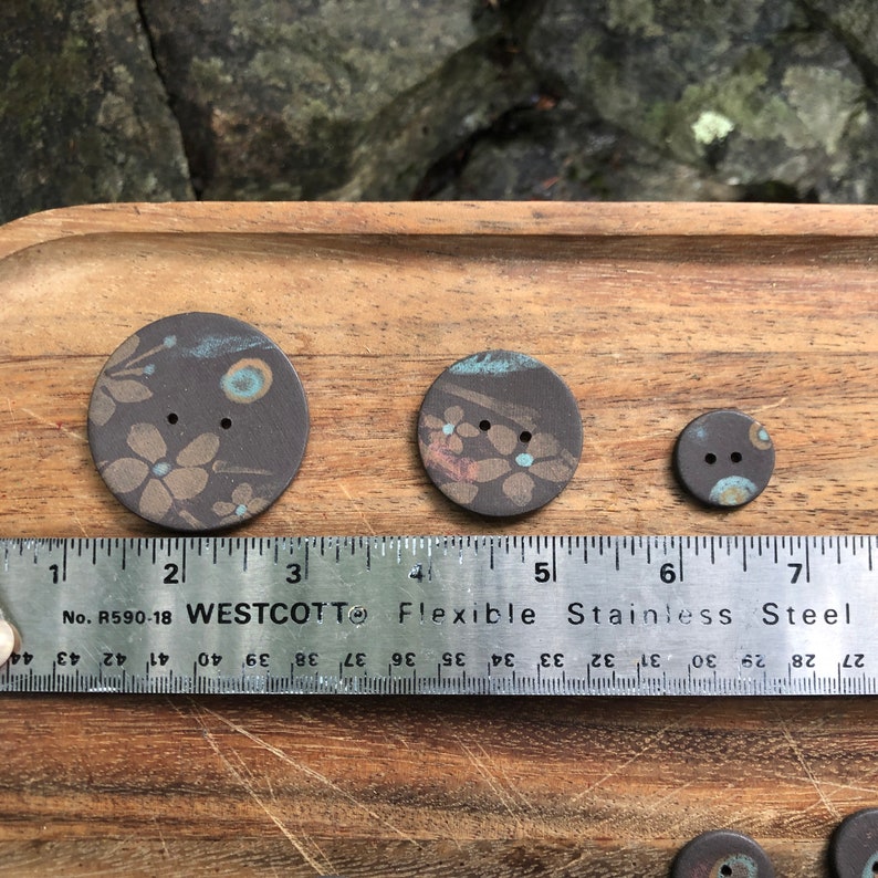 May include: Three ceramic buttons with a brown background and a floral pattern in shades of brown, pink, and blue. The buttons are arranged in order of size, with the largest button measuring approximately 1.5 inches in diameter.