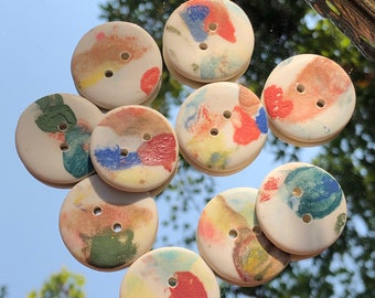 Könnte beinhalten: Neun bunte Keramikknöpfe mit jeweils zwei Löchern. Die Knöpfe sind mit abstrakten Mustern in Rot-, Blau-, Gelb-, Grün- und Weißtönen bemalt.
