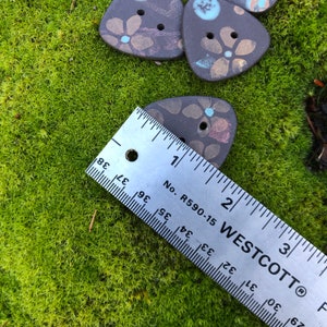 May include: A close-up of a ruler measuring a brown ceramic triangle button with a floral design. The button is approximately 1 inch in size. The ruler is marked with inches and fractions of an inch.