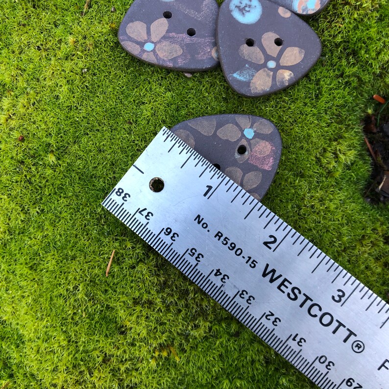 May include: A close-up of a ruler measuring a brown ceramic triangle button with a floral design. The button is approximately 1 inch in size. The ruler is marked with inches and fractions of an inch.