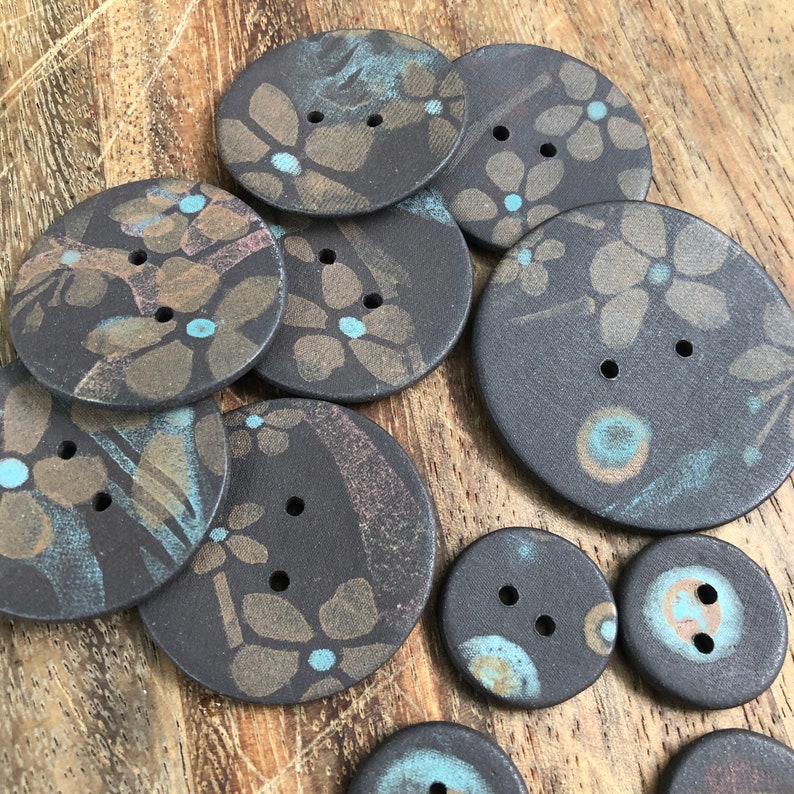 May include: A collection of 10 ceramic buttons with a black background and a floral pattern in shades of brown, gold, and blue.
