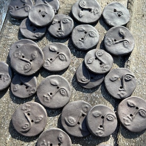 Peut inclure: Ensemble de boutons en céramique gris foncé, ornés de visages sculptés et de motifs de feuilles. Chaque bouton rond, percé de deux trous, présente des dimensions variables, pour un aspect artisanal. Idéal pour les projets créatifs.