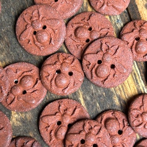 May include: A collection of terracotta-colored ceramic buttons with a bee design. The buttons are round and have a raised bee design with a textured surface.