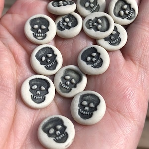 Puede incluir: Una colección de pequeños amuletos de calavera de cerámica blanca con detalles pintados en negro. Las calaveras están dispuestas en una mano, algunas superpuestas.