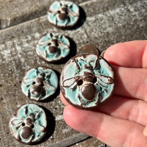 May include: Five ceramic buttons featuring a bee design. The buttons are brown and teal with a white accent. The bee is brown with a white stripe.