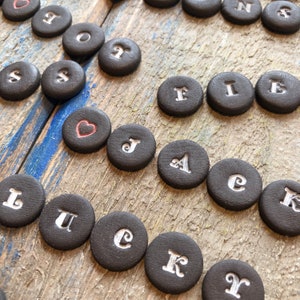 Op de afbeelding: Zwarte keramische tegels met witte letters die het woord "LUCK" spellen en een rode harttegel. De tegels zijn willekeurig gerangschikt op een houten oppervlak.