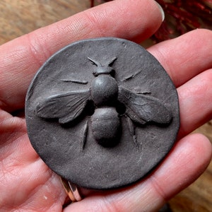 May include: A dark brown clay button featuring a raised bee design. The bee is detailed with wings, legs, and a stinger.