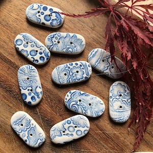 May include: A selection of oval ceramic buttons with a white and blue floral pattern. The buttons, each with two holes, are arranged on a wooden surface. The designs include dots and stylized flowers. The buttons are approximately 3 cm in length.