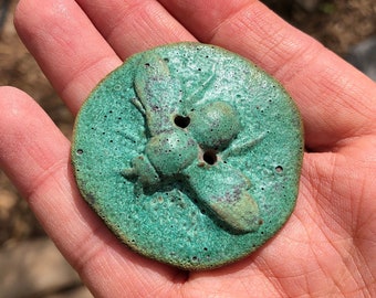 May include: A circular, turquoise button featuring a raised bee motif. The button has two holes and a textured surface. The bee design includes wings and body segments. The button is likely made of ceramic or metal.
