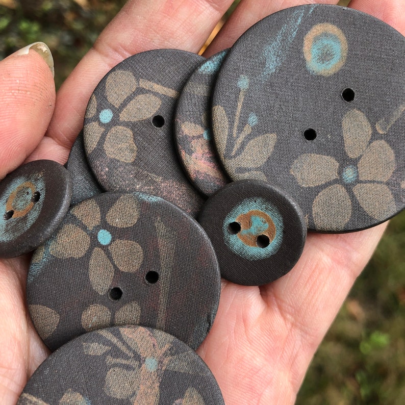 May include: A collection of six dark brown ceramic buttons with floral patterns and blue and gold accents. The buttons are arranged in a hand.