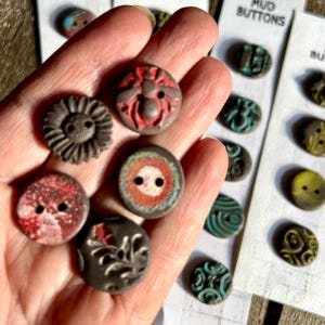 May include: A hand holding six ceramic buttons with various designs. The buttons are in shades of brown, red, and blue. The buttons have two holes each. The buttons are on a white background with the text "MUD BUTTONS".