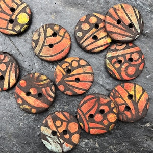 Op de afbeelding: Een verzameling van tien kleurrijke keramische knopen met verschillende patronen en ontwerpen. De knopen zijn rond en hebben twee gaten. De kleuren zijn oranje, geel, bruin en zwart.