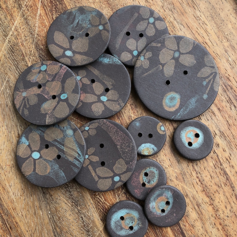 May include: A collection of 11 round ceramic buttons with various patterns and colors. The buttons feature a mix of blue, brown, and white colors with floral and abstract designs.