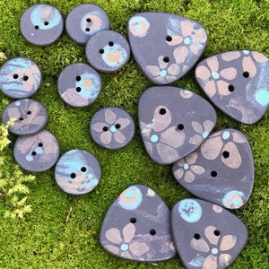 May include: A collection of handmade ceramic buttons with floral patterns in shades of black, white, and blue. The buttons are various shapes and sizes, including round, triangular, and oval.