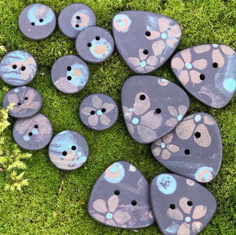 May include: A collection of handmade ceramic buttons with floral patterns in shades of black, white, and blue. The buttons are various shapes and sizes, including round, triangular, and oval.