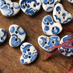 May include: A collection of heart-shaped ceramic buttons with a blue and white floral pattern. The buttons have two holes each and are arranged on a wooden surface.