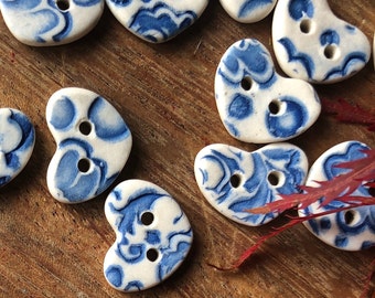 May include: A collection of heart-shaped ceramic buttons with a blue and white floral pattern. The buttons have two holes each and are arranged on a wooden surface.