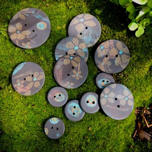 May include: A collection of 11 ceramic buttons with floral designs in shades of blue, pink, and brown. The buttons are arranged on a bed of green moss.