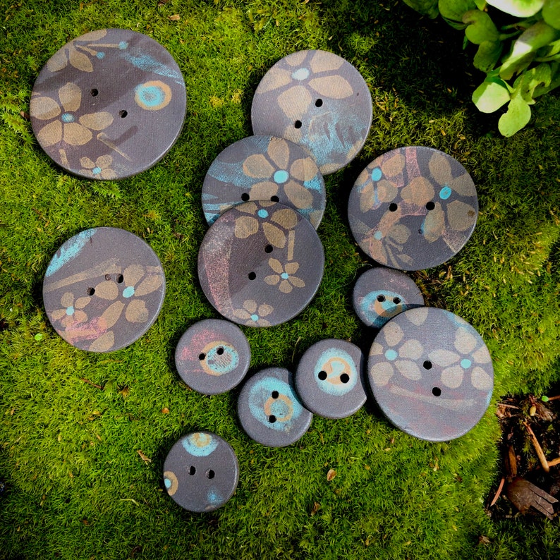 May include: A collection of 11 ceramic buttons with floral designs in shades of blue, pink, and brown. The buttons are arranged on a bed of green moss.