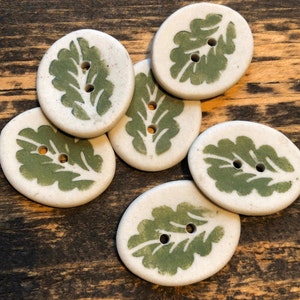 May include: Six white ceramic buttons with green leaf designs. The buttons are oval shaped and have two holes.