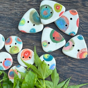 Puede incluir: Surtido de botones de cerámica coloridos con fondo blanco y dos agujeros. Los botones son de varias formas, incluyendo círculos y triángulos. Algunos botones tienen un patrón de círculos coloridos.