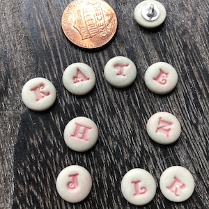 May include: Nine white ceramic buttons with pink letters spelling out the word "KATHLEEN".