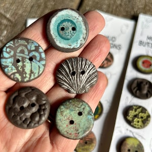May include: A collection of handmade ceramic buttons featuring a variety of designs and colours, including a blue and green button with a circular pattern, a black and white button with a seashell design, a brown button with a textured surface, and a green and brown button with a speckled design. The buttons are displayed on a white background with the text "MOLD BUTTONS".