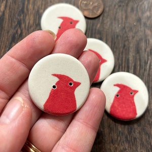 May include: Four ceramic buttons featuring a red cardinal design. The buttons are round and have a white background.