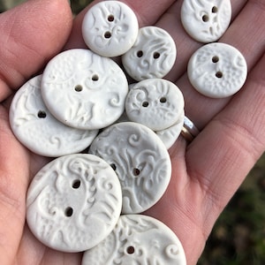 May include: A collection of white ceramic buttons with intricate floral and swirl designs. The buttons are various sizes and have two holes each.