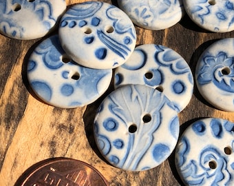 May include: A collection of 10 blue and white ceramic buttons with various floral and geometric patterns. The buttons are round and have two holes each. The buttons are arranged on a wooden surface.