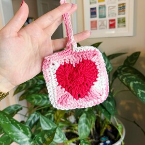 May include: A handmade crochet square with a red heart in the center, surrounded by pink and white. The square is attached to a pink loop. The item is held in front of a houseplant.