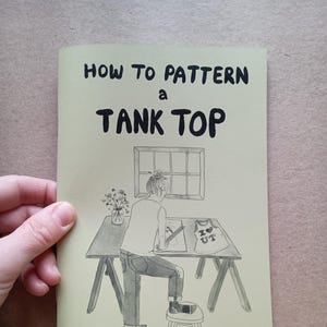 May include: A soft green book titled "HOW TO PATTERN A TANK TOP" with an illustration of a person designing a tank top. The author's name, Louisa Owen Sonstroem, is printed at the bottom. The book is held by a hand.