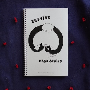 May include: A spiral-bound notebook with the words "FESTIVE HAND SEWING" in black lettering. The cover features a black and white illustration of a person sewing. The notebook rests on a dark blue fabric with red embroidered accents.