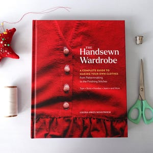 May include: A red book titled "The Handsewn Wardrobe" with sewing tools. The book cover features a close-up of a red garment with buttons. Other items include a star-shaped pin cushion, scissors, a thimble, and thread.