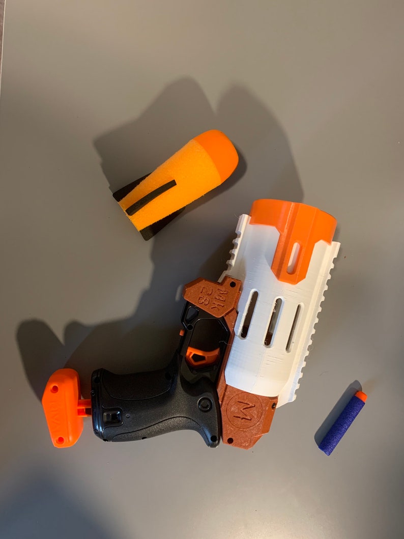 K&ouml;nnte beinhalten: Eine wei&szlig;e und orangefarbene Spielzeugpistole mit schwarzem Griff und einem blauen Dart. Die Pistole hat einen braunen Lauf und ein wei&szlig;es Magazin. Die Pistole ist mit "MARK 32" beschriftet.
