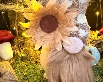 Puede incluir: Una muñeca de gnomo hecha a mano con barba de piel sintética marrón, nariz rosa y un acento de girasol. El gnomo lleva un sombrero con estampado marrón y blanco y un puño a rayas azules y blancas. La muñeca está sobre un lecho de musgo verde.