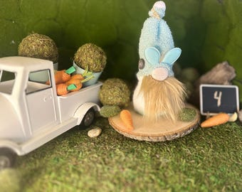 Puede incluir: Una escena de Pascua con un camión de juguete blanco que transporta zanahorias y bolas de musgo. Un gnomo con un sombrero azul y orejas de conejo está sobre una rodaja de madera. Una pequeña pizarra muestra el número 4.